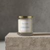 A glass jar candle with a gold metal lid and a cream label reading 'Pumpkin Pie Spice' sits on a textured stone pedestal against a gray concrete wall.