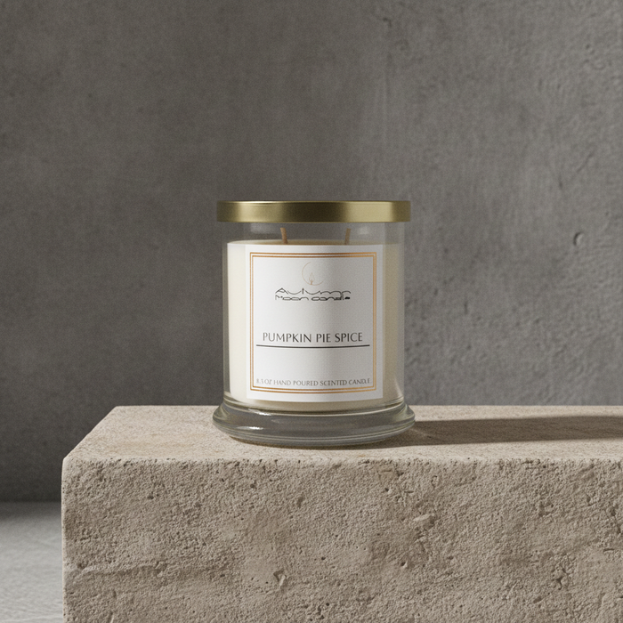 A glass jar candle with a gold metal lid and a cream label reading 'Pumpkin Pie Spice' sits on a textured stone pedestal against a gray concrete wall.